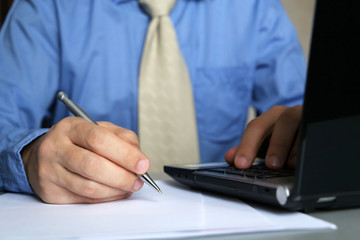 Fototapeta premium Man in blue shirt and tie works in office, sitting at a table with laptop and pen in hand. Male hands close-up, writing hand, concept of clerk, manager, businessman, official or bank employee