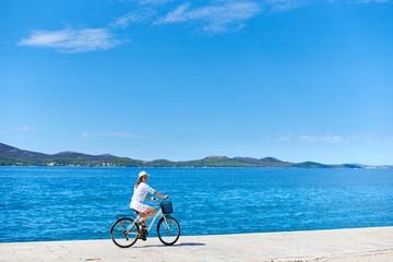Happy woman in white summer closing and hat riding a bicycle along stony sidewalk on blue sparkling sea water and resort town at foot of mountains on opposite shore background. Tourism and vacations.