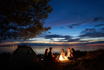 Night summer camping on sea shore. Group of young tourists sitting, laughing in high grass around...