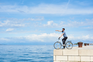 Fototapeta premium Young bearded man on bicycle on high paved stone sidewalk pointing at distant mountain on opposite shore on clear blue sea water and small sailing boat background. Active holiday and tourism concept.