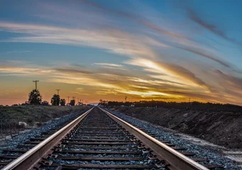 Poster Parallelle spoorlijnen die langs landbouwvelden gaan en de kleurrijke zonsondergang in gaan © motionshooter