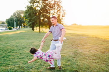 Naklejka na ściany i meble Handsome hipster father with mustache, hair style whirling his little daughter ootdoor on green field in park. Dad playing carousel with his cute child in multicolored dress. Parent with kid enjoying