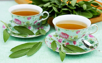 Sage herbal tea served in minty with pink roses china porcelain cups standing on linen tablecloth with bunch of sage herb in a wooden tray on the background. Alternative medicine concept