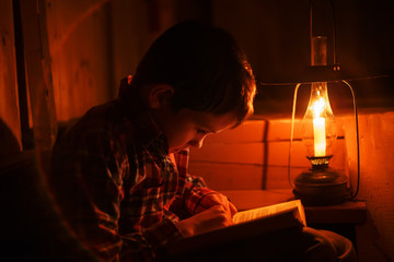  child sitting on a dark staircase with a book at night. boy reading in the light of an old lamp