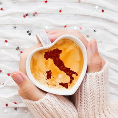 Woman holds a cup of hot coffee with cinnamon silhouette of Italy. Winter wanderlust and Valentine's Day fabric background with sparkling silver and red confetti.
