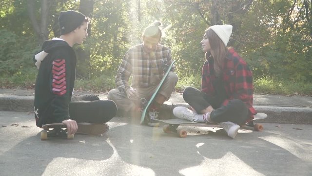 Young people are sitting with skateboards next to each other outdoors. Hipster friends chatting while sitting with a skateboard in the park. Skateboarder guy in a plaid shirt adjusts his skateboard.