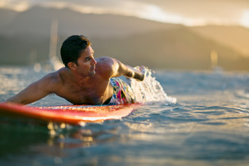 Male surfer lies on his board,  looks over his shoulder and paddles in anticipation of a coming wave.
