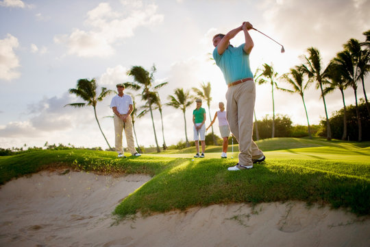 Man and two women watch a male golfer take a shot over a sand trap.