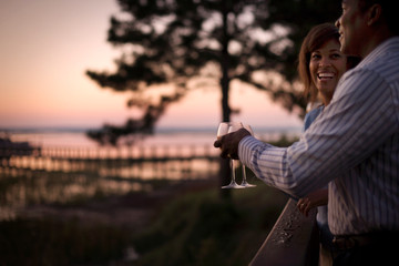 Laughing couple toasting wine glasses on a balcony at sunset.