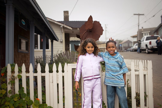 Portrait of a young boy and his young sister standing beside a white picket fence in a suburban street.