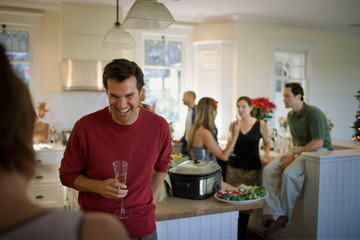Laughing young man holding a champagne flute while inside a kitchen with friends.