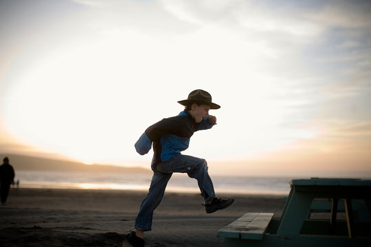 Young boy stepping onto a picnic table at the beach while wearing a hat.