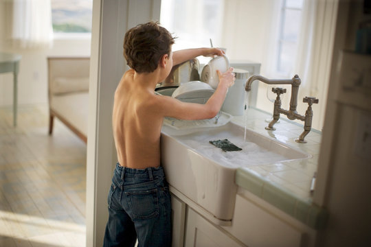 Shirtless young boy washing dishes in the kitchen sink.