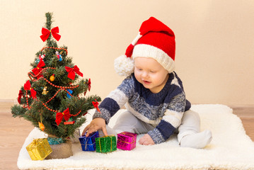 Funny baby in Santa Claus hat sitting near the Christmas tree with gifts
