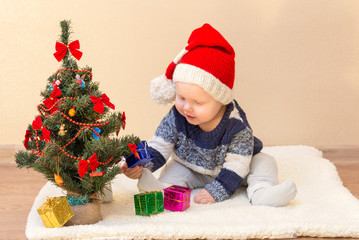 Funny baby in Santa Claus hat sitting near the Christmas tree with gifts