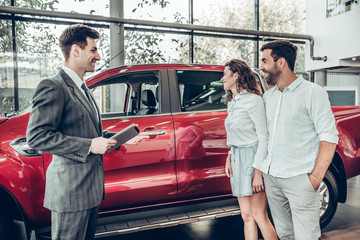 Attractive couple is talking to car sales manager in luxury car dealership and looking at beautiful...