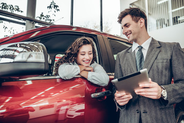 Beautiful young woman is talking to handsome sales manager while choosing a car in dealership.