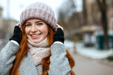 Closeup shot of cute ginger woman with long hair wearing knitted cap and scarf walking at the city. Empty space