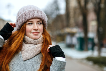 Closeup shot of elegant ginger girl with long hair wearing knitted cap and scarf walking at the city. Empty space