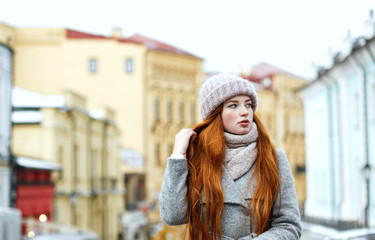 Street portrait of glorious redhead woman with long hair wearing warm winter apparel posing at the street. Empty space