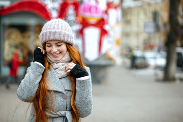 Urban portrait of adorable ginger girl with long hair wearing warm clothes walking at the city. Empty space