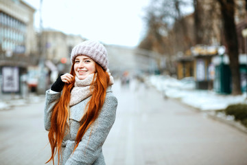 Urban portrait of positive ginger model with long hair wearing warm clothes walking at the city. Empty space