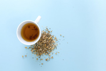 Cup of freshly brewed chamomile tea with heap of dried plant, on blue background.