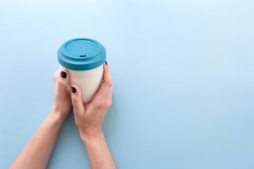 Hands holding bamboo reusable takeaway cup with lid on, on blue background.