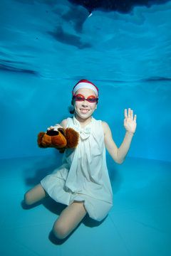 A child with a toy-dog in his hand sitting at the bottom of the pool under the water in a white dress and swimming goggles, smiling and waving on a blue background. Vertical orientation of the image