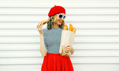 Pretty smiling young woman wearing red beret holding croissant, paper bag with a long white bread...