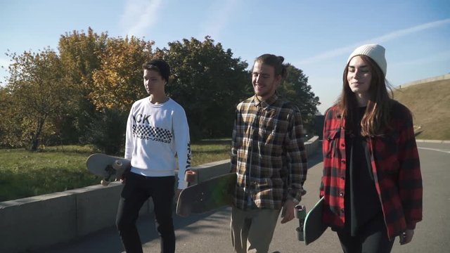 Teenagers with skateboards walk outdoors. Two guys and a girl spend time with skateboards on their hands in the park. Slow motion. Hobbies and lifestyle.