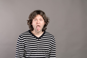 Portrait of funny shaggy crazy handsome man in striped sweater looking at camera with big open mouth and tongue out, teases. Wide opened eyes indoor studio shot, isolated on gray background.