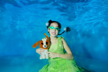 A little girl swims and poses underwater in a green dress and swimming glasses at the bottom of the pool, holds a toy - dog in her hands, looks at the camera and smiles. Portrait. Shooting underwater