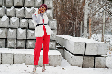 African american woman in red pants and white fur coat jacket posed at winter day against snowy stone background.