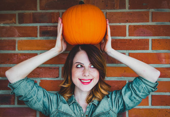 Redhead woman in jeans clothes holding orange autumn pumpkin. Portrait on brick background. Vegetable may be ECO product