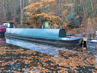 a scenic view of narrow boats in a canal surrounded by golden fallen autumn leaves with reflections on the water and forest trees in hebden bridge west yorkshire