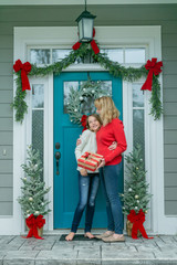 Obraz na płótnie Canvas Mother and Daughter with a Christmas Gift Outside Their Home with a Festive Decorated Front Door