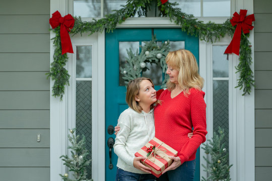 Mother And Daughter With A Christmas Gift Outside Their Home With A Festive Decorated Front Door