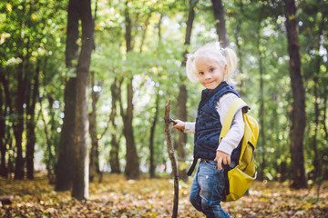 Theme outdoor activities in nature. Funny little Caucasian blonde girl walks walks hiking in the forest on rough terrain with a large backpack. Uses walking stick