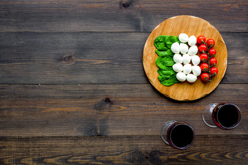 Italian cuisine, food concept. Italian flag made of mozzarella, tomatoes, basil on wooden cutting board near glass of red wine on dark wooden background top view copy space