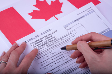 Filling Application (female hands and wood pen) form for Canadian Citizenship - Adults. Immigration, Refugees and Citizenship Canada on Canadian flag surface. 