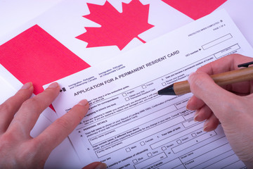 Filling Application (female hands and wood pen) form for Canadian Citizenship - Adults. Immigration, Refugees and Citizenship Canada on Canadian flag surface.