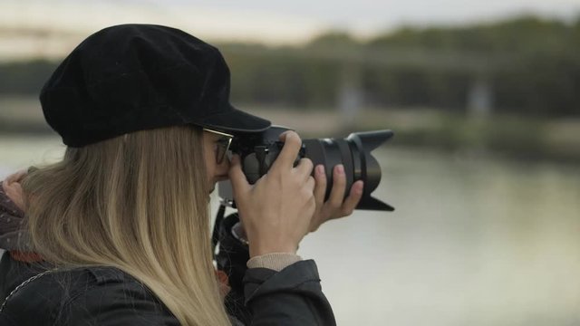 Professional young blonde woman photographer with digital camera taking pictures of autumn river at evening city city. Close Up, shallow depth of field.