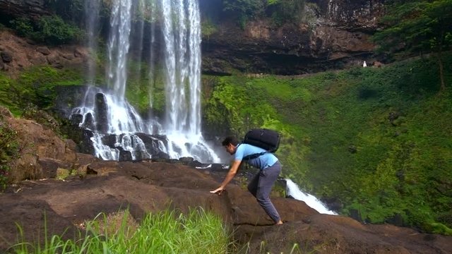 barefoot guy with backpack climbs up stone by waterfall