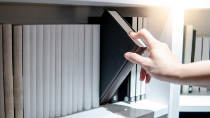 Male hand choosing and picking black book from white bookshelf in public library. Education...