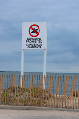 Large Swimming Prohibited Dangerous Currents warning sign at an ocean beach shoreline with a wooden fence along the sand dunes along the water edge.
