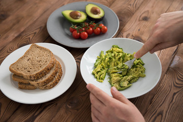 Hands of cook chef preparing healthy avocado sandwich on dark rye toast bread made with fresh avocado paste, cherry tomatoes on brown wooden background