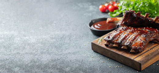 Closeup of pork ribs grilled with BBQ sauce and caramelized in honey. Tasty snack to beer on a wooden Board for filing on dark concrete background. With copy space - obrazy, fototapety, plakaty