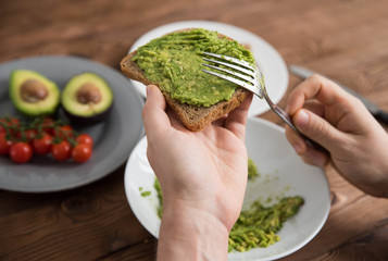 Hands of cook chef preparing healthy avocado sandwich on dark rye toast bread made with fresh avocado paste, cherry tomatoes on brown wooden background