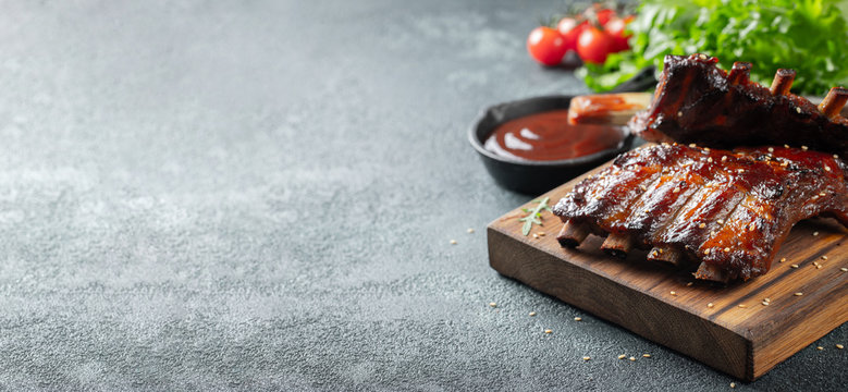 Closeup of pork ribs grilled with BBQ sauce and caramelized in honey. Tasty snack to beer on a wooden Board for filing on dark concrete background. With copy space